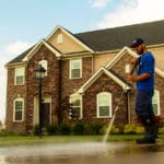 Power washing technician applying siloxane sealer on concrete in front of brick and vinyl home