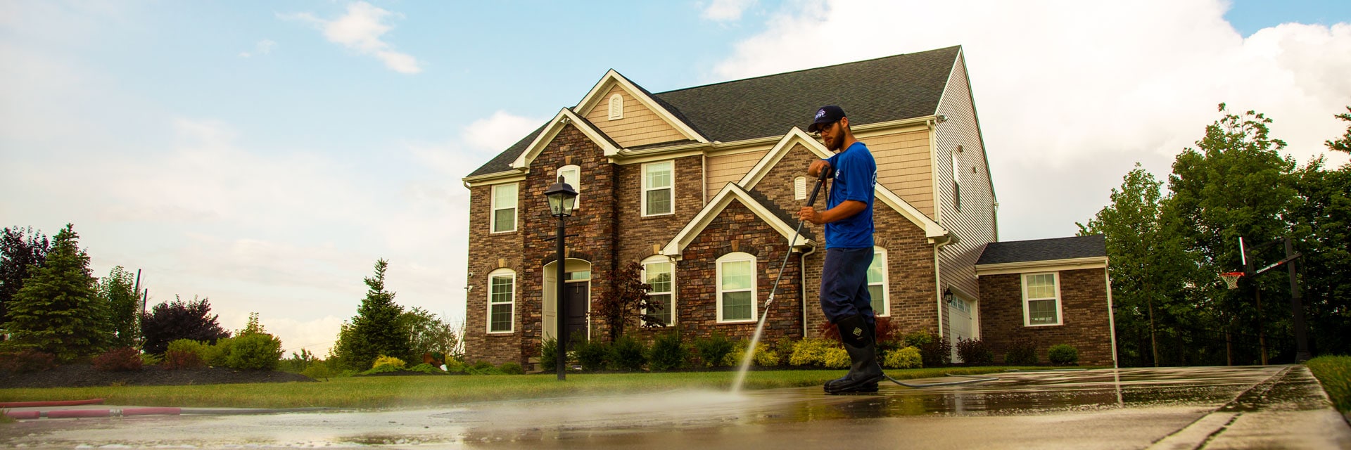 Power washing technician applying siloxane sealer on concrete in front of brick and vinyl home