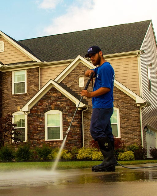 Power washing technician applying concrete sealer on concrete in front of brick and vinyl home